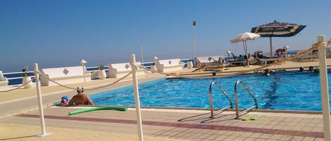 Ein Poolbereich mit freiem Blick auf das Meer, Liegestühle, Sonnenschirme und ein Rettungsschwimmerstand.