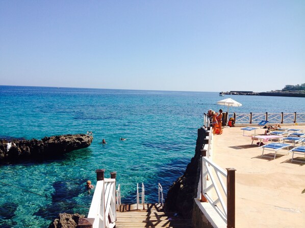 A pier leading into clear blue waters with a sandy beach area and lounge chairs. - La Marsa Vacances (Palermo)