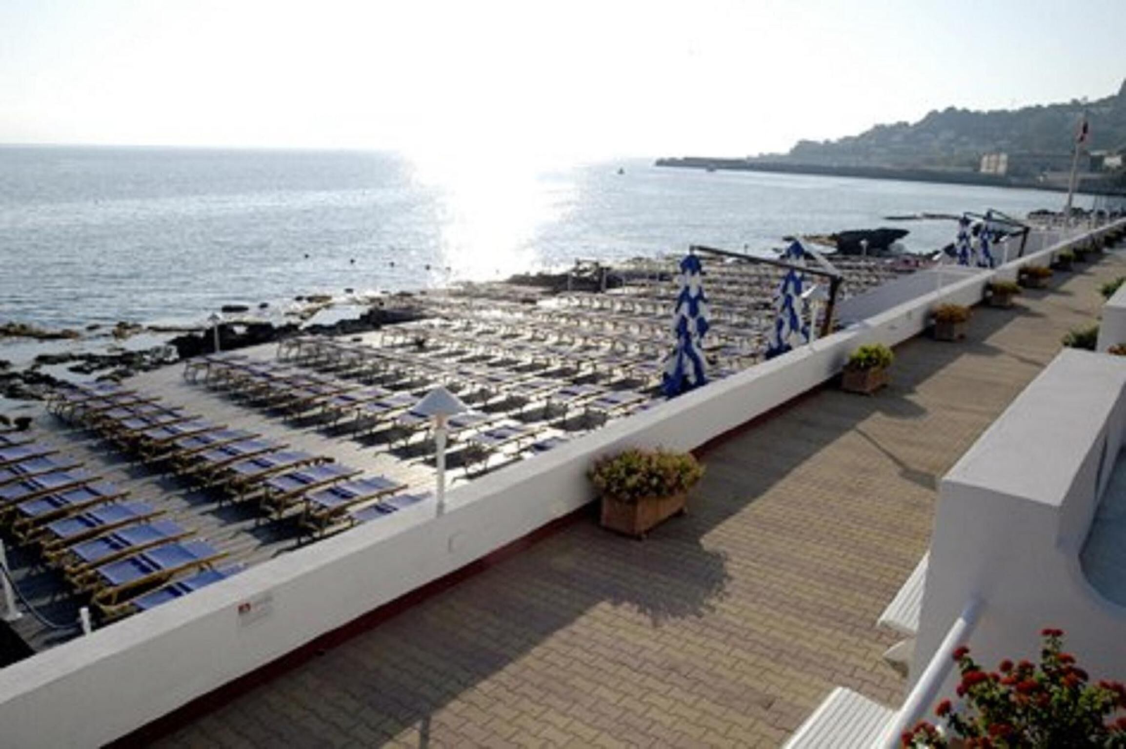 A seaside terrace with rows of sun loungers and umbrellas, a paved walkway, and a view of the ocean.