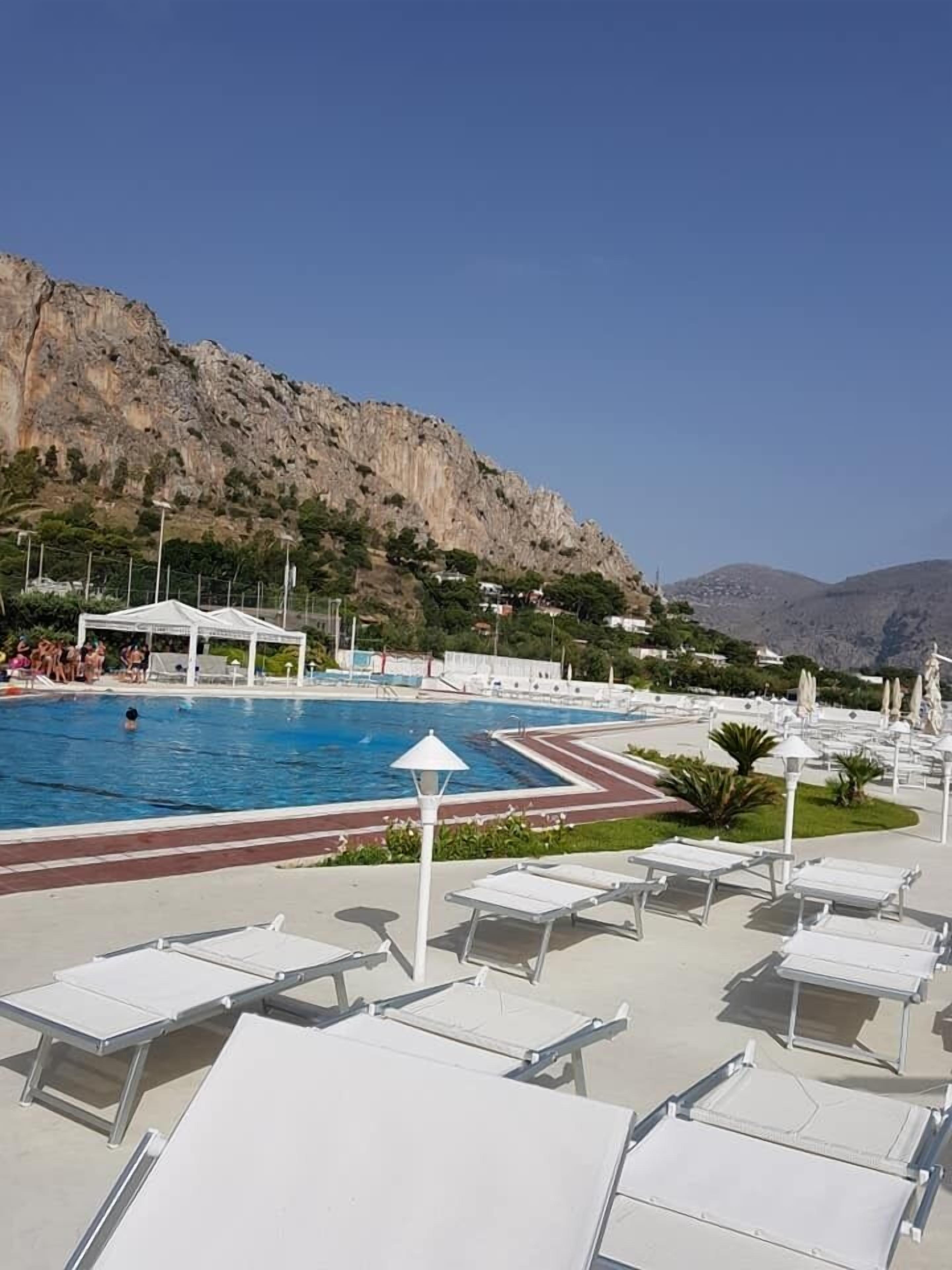 Zona de piscina con sillas de descanso y sombrillas, rodeada por un paisaje montañoso.