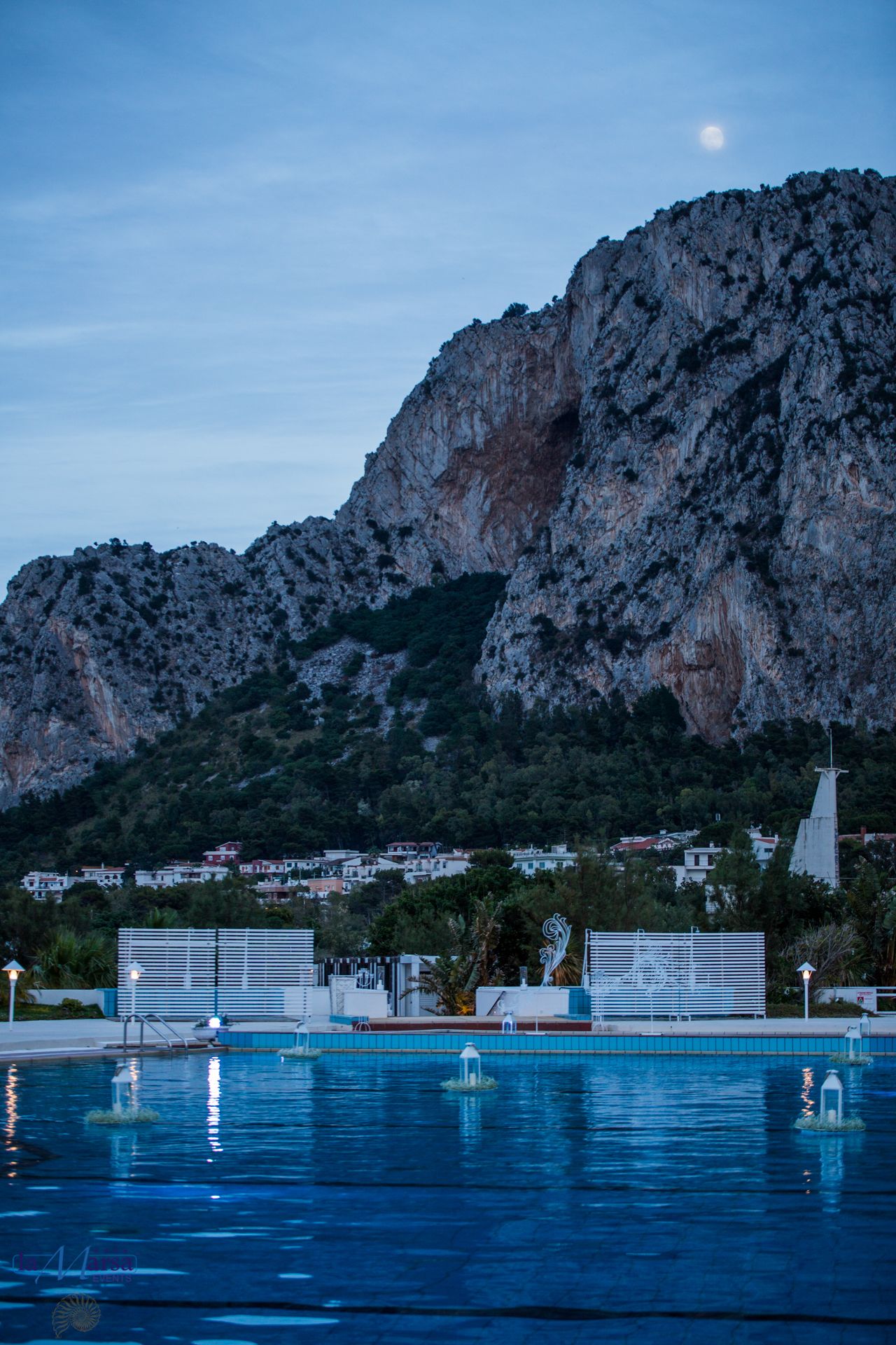 Una piscina con montañas al fondo, un edificio blanco con techo azul y una aguja de iglesia.