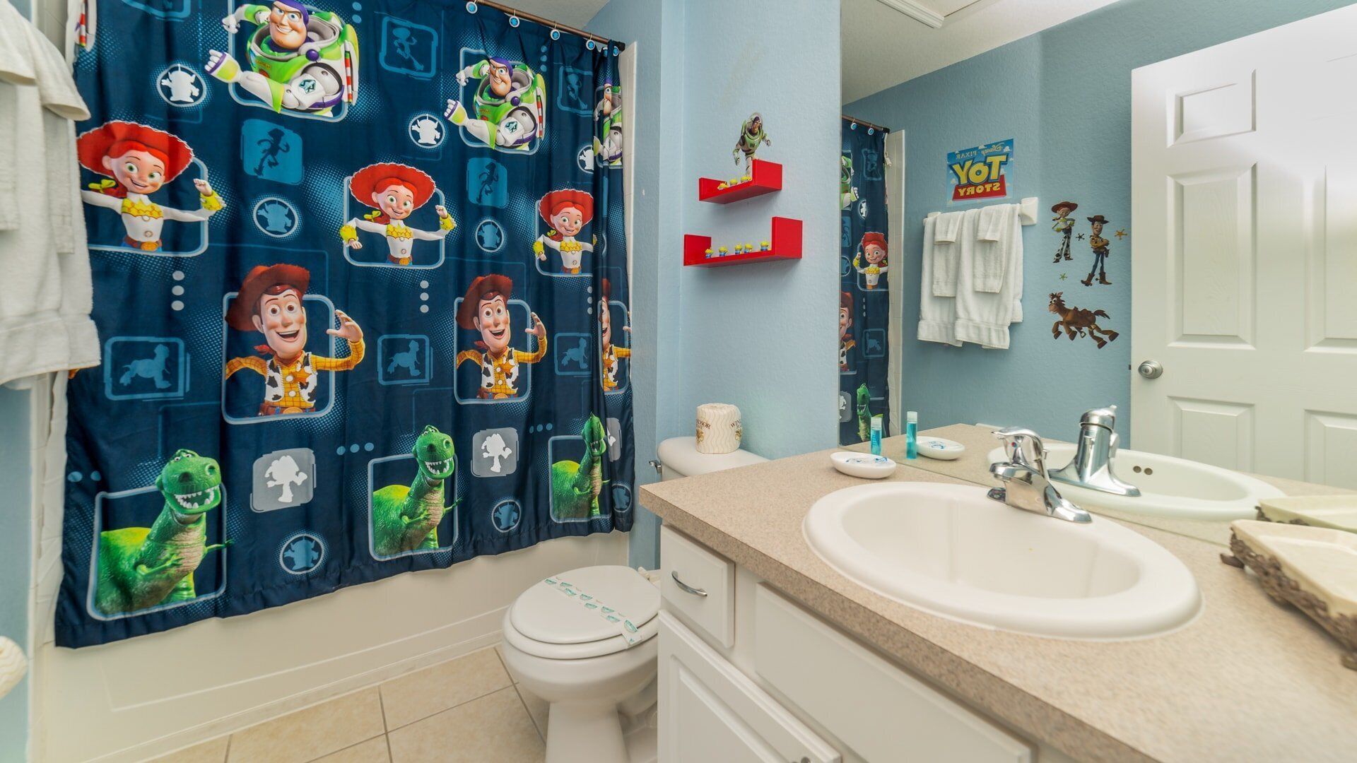 A bathroom with a blue shower curtain featuring cartoon characters, a white toilet, a sink with a mirror, and a towel rack.