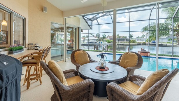 Comedor con mobiliario de mimbre, una mesa redonda y vistas a una piscina y edificios.
