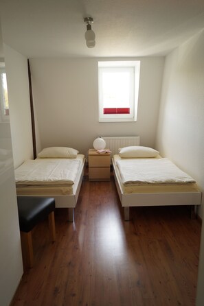 A small, clean room with two single beds, a wooden bench, a nightstand with a lamp, and a window with blinds.