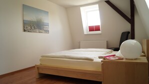 A bedroom with a wooden bed, a window with blinds, a radiator, a desk with a lamp, and a painting on the wall. - Strandleben Fehmarn - Ferienwohnung Nord (Fehmarn)