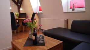 A cozy living room with a wooden coffee table, a black sofa, a dining area with a table and chairs, and a window with red blinds. - Strandleben Fehmarn - Ferienwohnung Nord (Fehmarn)