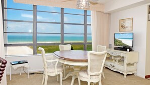 A room with a large window overlooking a beach, a dining table with chairs, a television, and a small table with a laptop.