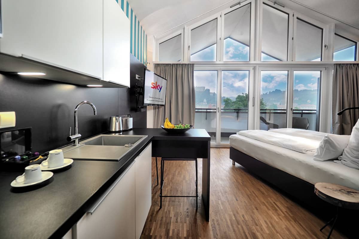 Habitación de hotel moderna con cocina, una mesa de comedor, sofá y ventanas amplias con vista a la ciudad.