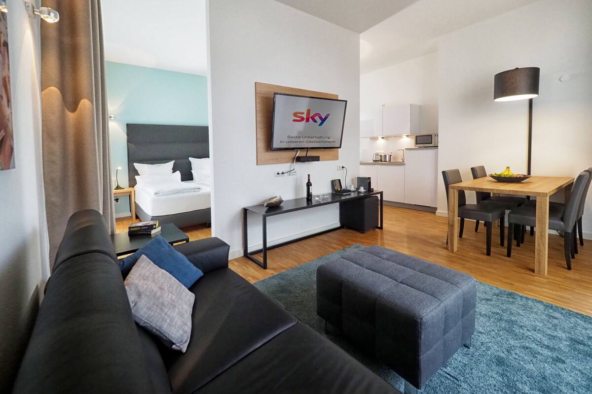 Una habitación de hotel moderna con zona de estar, una mesa de comedor y un televisor en la pared.