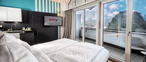 Quarto de hotel com cama, televisão presa à parede, varanda com vista para a cidade e área de cozinha.