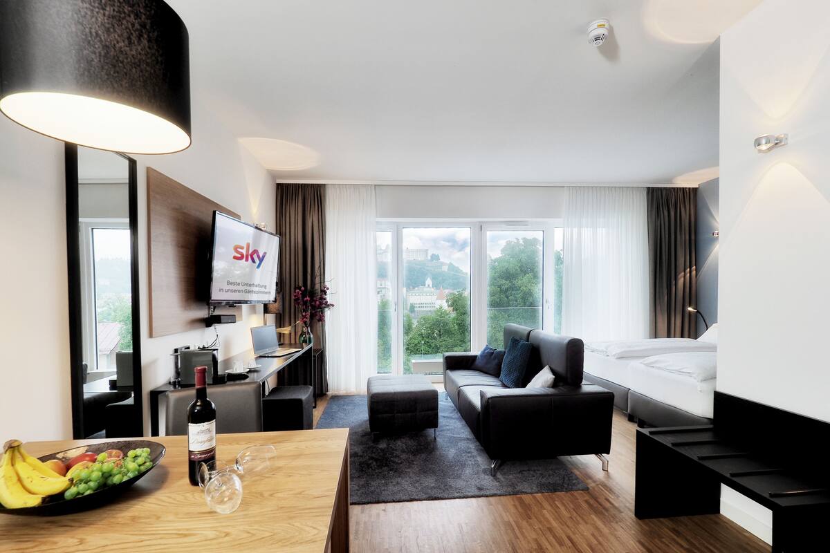 Una habitación de hotel moderna con un escritorio de madera, un sofá negro, una cama y un gran ventanal con vistas a la ciudad.