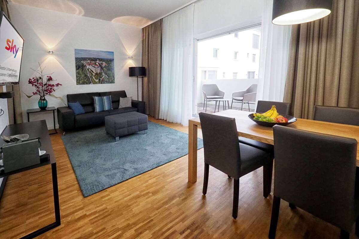 Una sala moderna con un sofá, un reposapiés y una mesa de comedor con sillas. Hay un cuadro en la pared, un florero con flores y una lámpara.