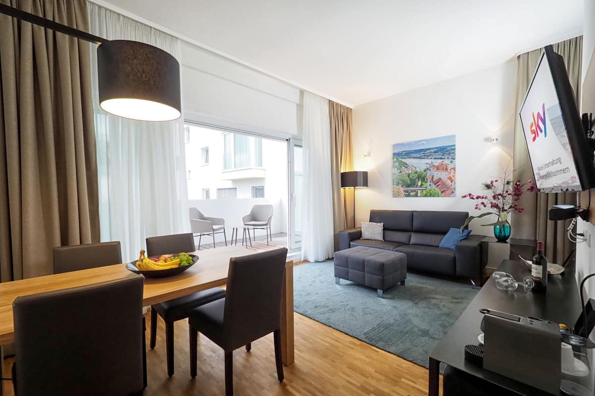 Una sala de estar moderna con comedor, un sofá, una mesa de centro y un televisor. Hay un balcón con vistas a edificios.