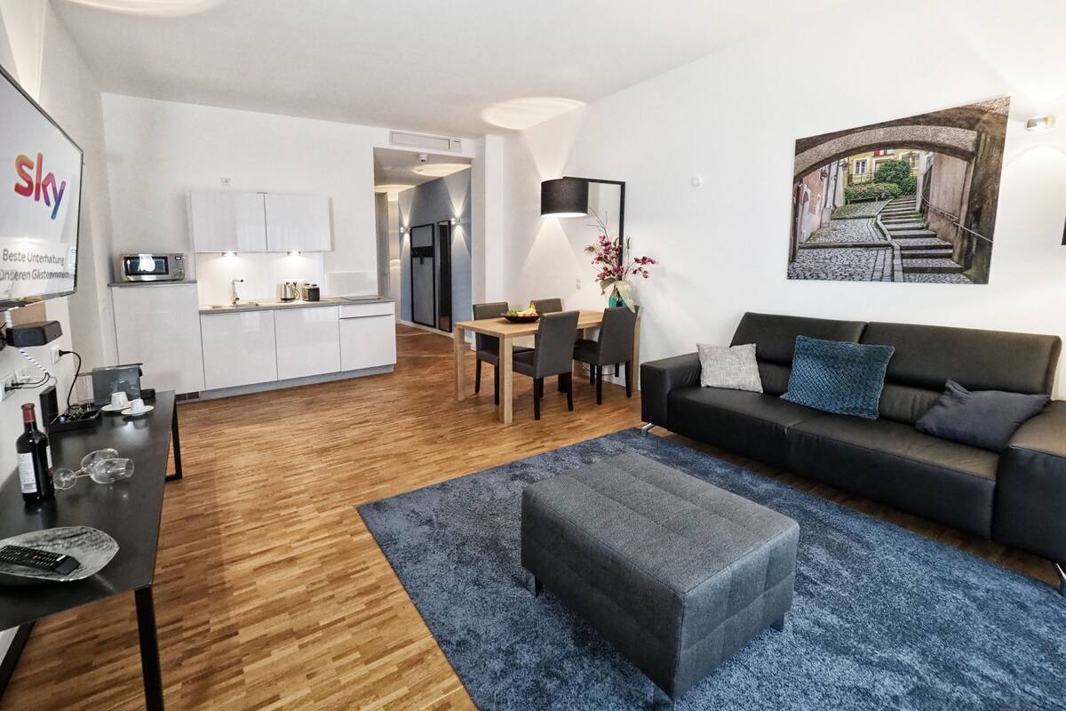 Una sala de estar moderna con un sofá oscuro, un tapete azul y una zona de comedor con mesa y sillas de madera.