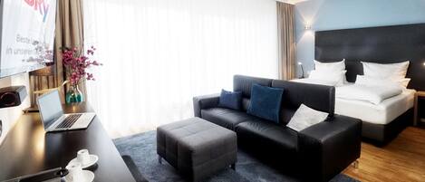 A modern hotel room with a dark leather sofa, a bed with white linens, a desk with a laptop, and a large window with sheer curtains.