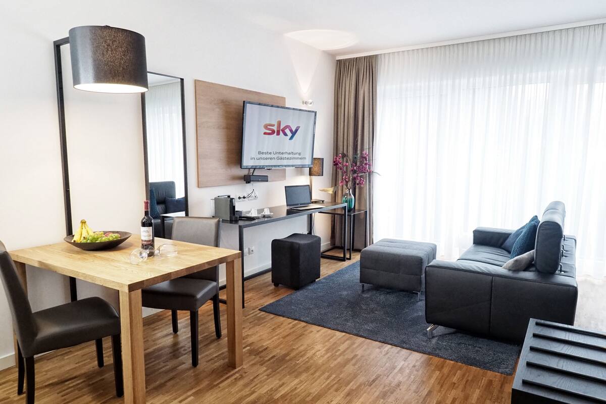 Una habitación de hotel moderna con zona de comedor, sofá, televisor y un gran ventanal con cortinas.