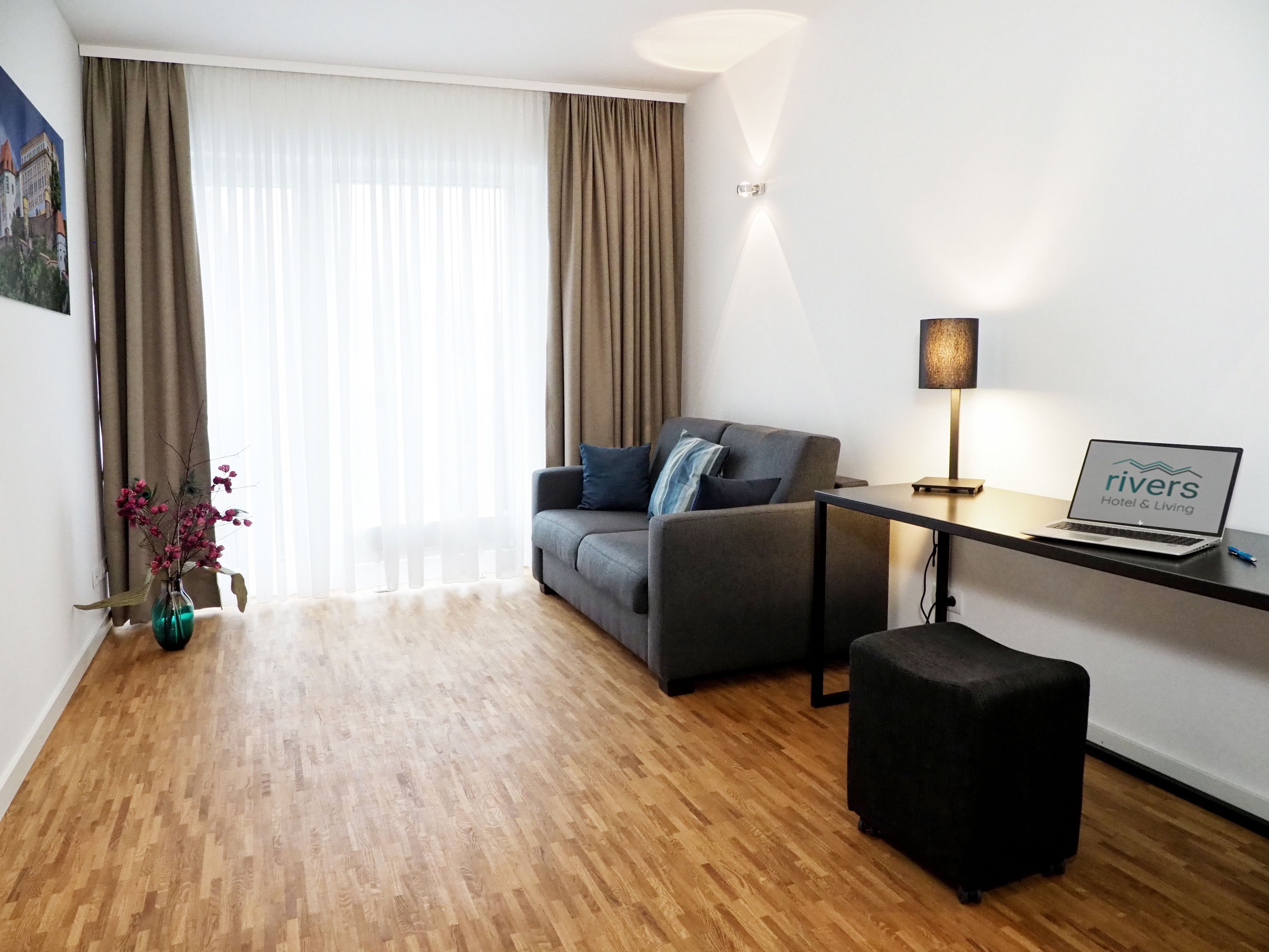 Una sala moderna con un sofá gris, un pequeño taburete negro, una mesa de centro de vidrio y una computadora portátil mostrando 'rivers Hotel & Living'.