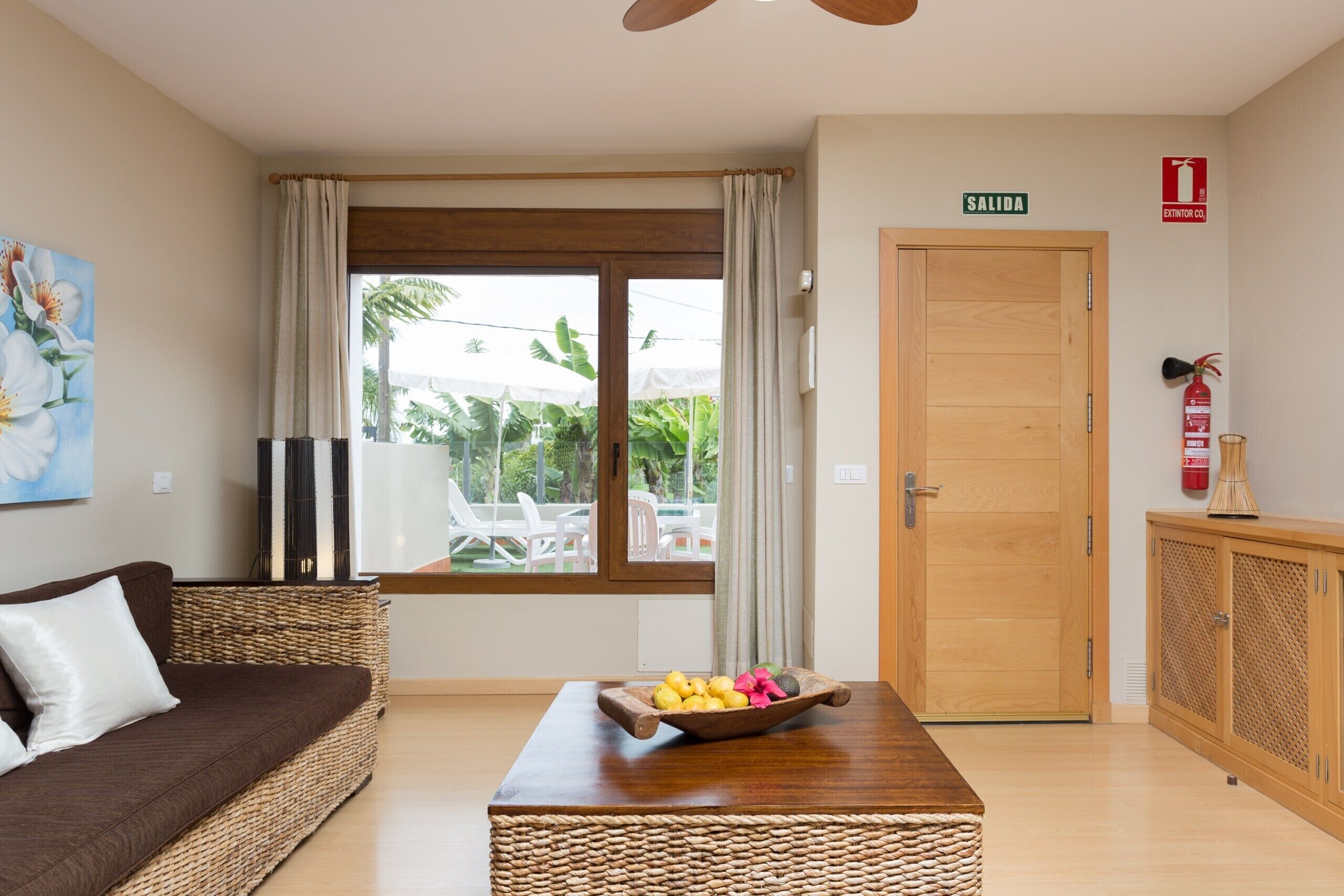 Una habitación con una puerta de madera que dice 'SALIDA', un sofá de mimbre, una mesa de madera con un frutero y vistas a muebles de exterior a través de una ventana.