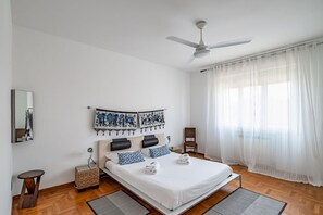 A bedroom with a bed, a ceiling fan, a window with sheer curtains, a wall-mounted mirror, and a small side table.