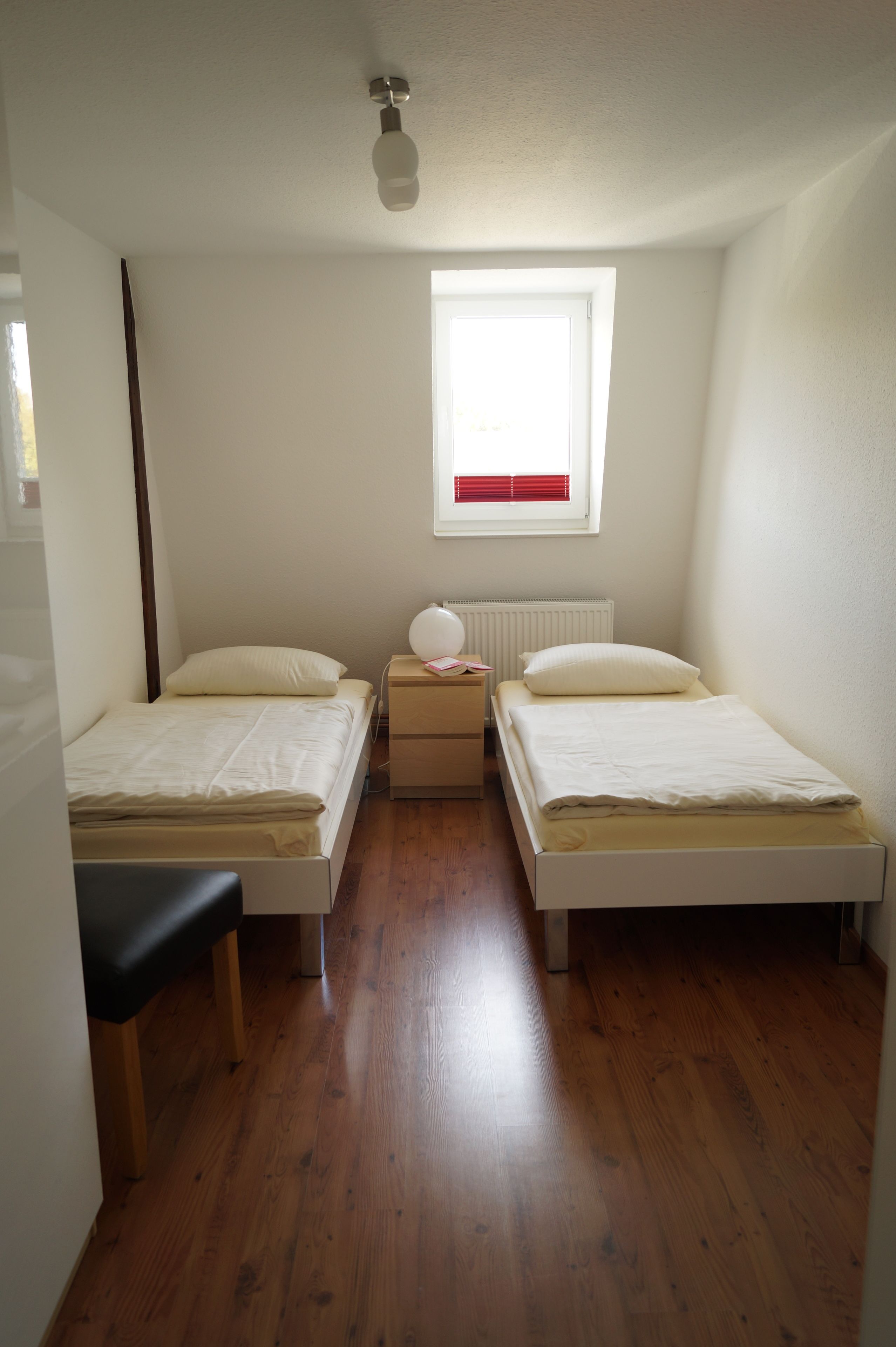 Une petite chambre propre, avec deux lits simples, un banc en bois, une table de chevet avec une lampe et une fenêtre avec des stores.