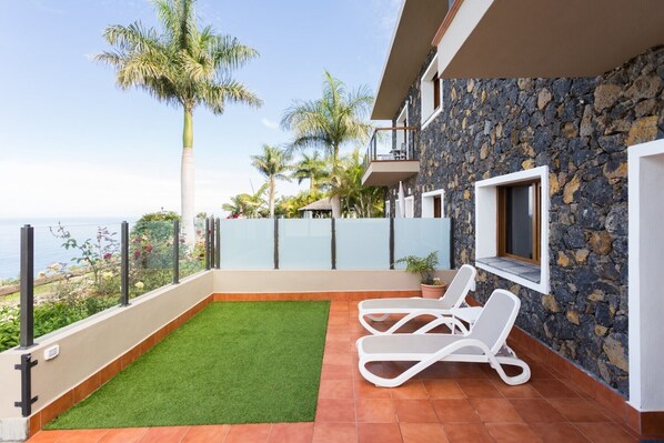 Ein Balkon mit künstlichem Rasen, weißen Liegestühlen, einer Steinmauer und Blick auf das Meer.