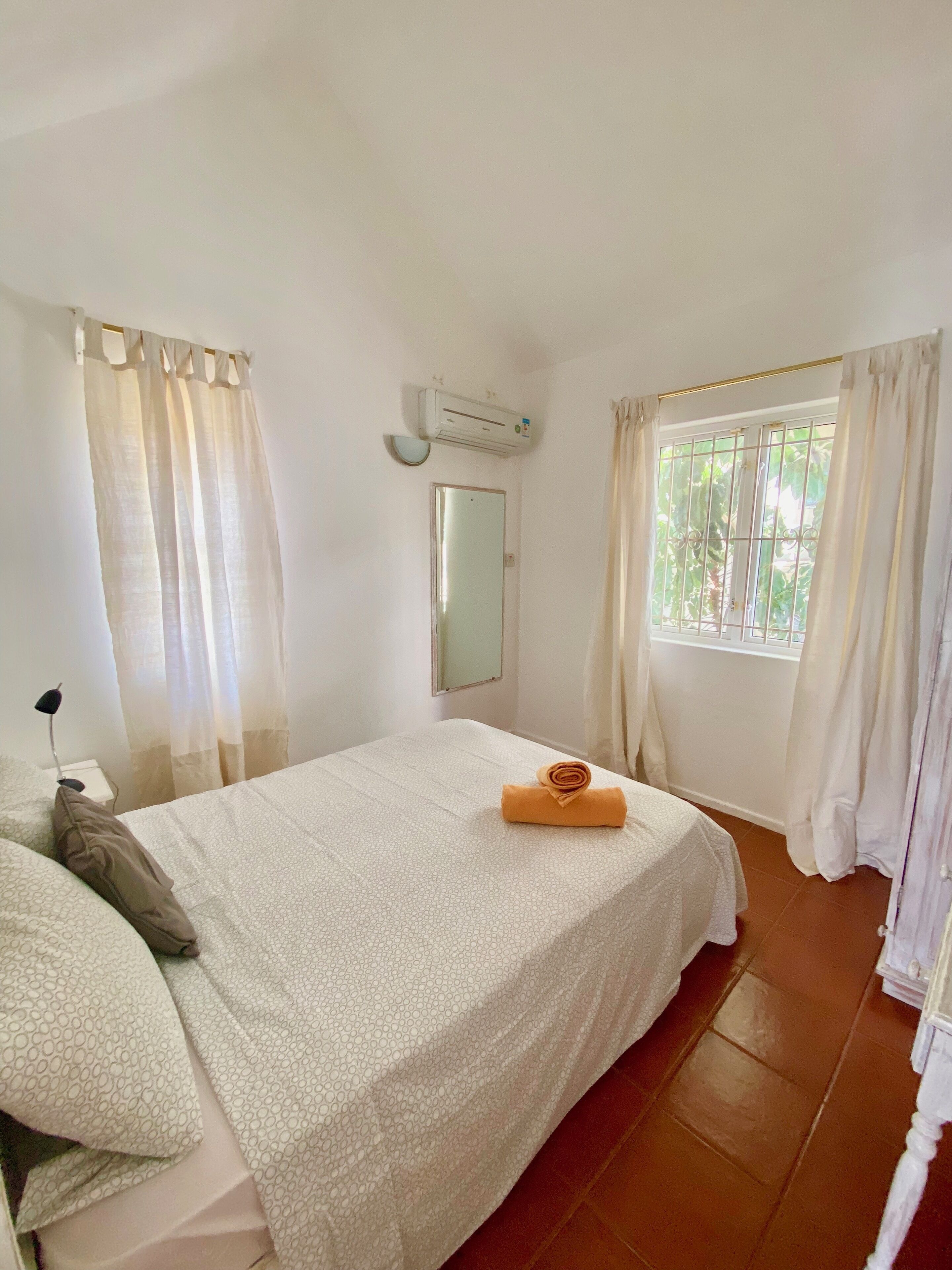 A bedroom with a bed, a mirror, an air conditioning unit, and a window with curtains.