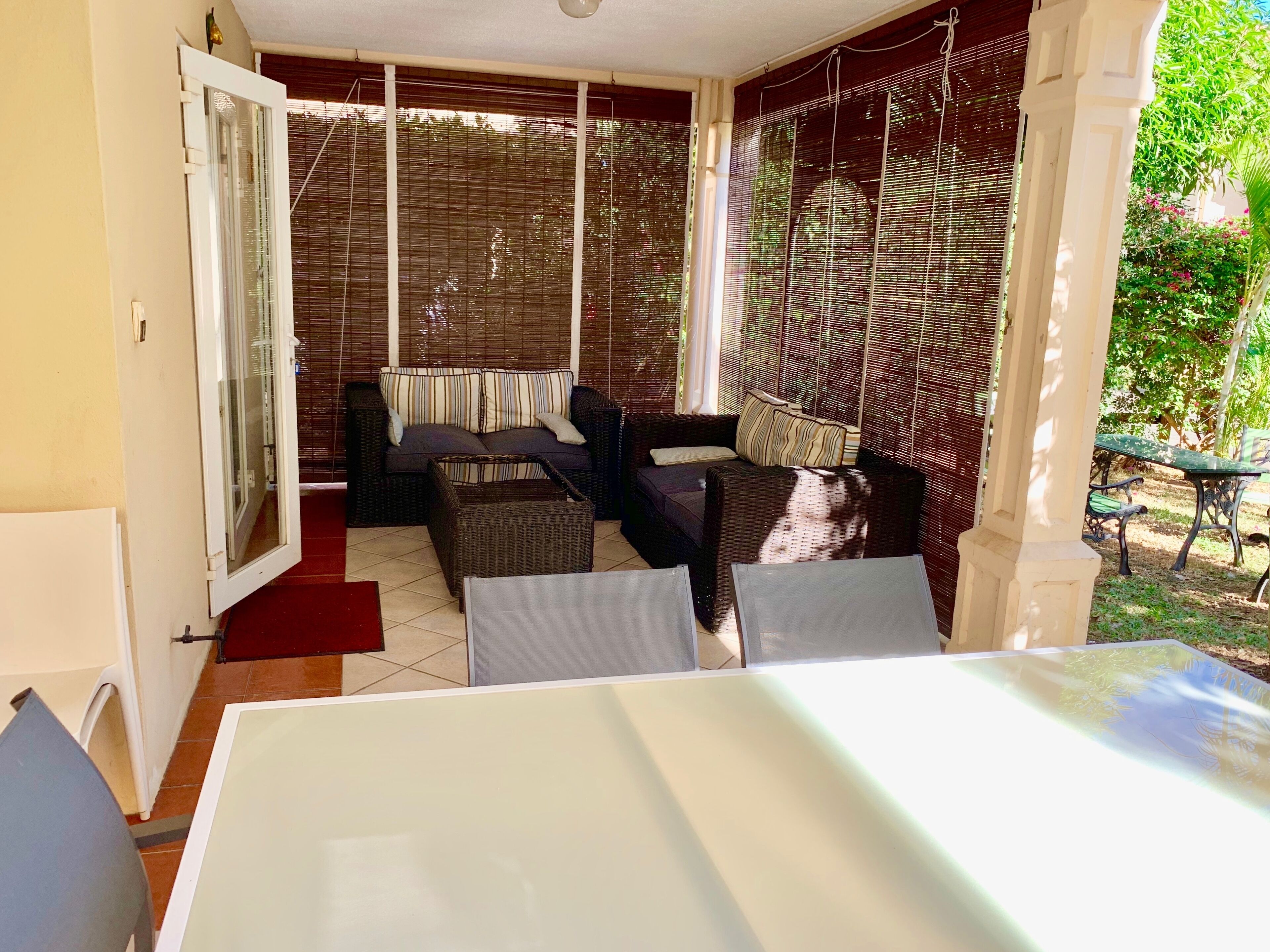 A covered outdoor seating area with wicker furniture, a white table, and a view of the garden.