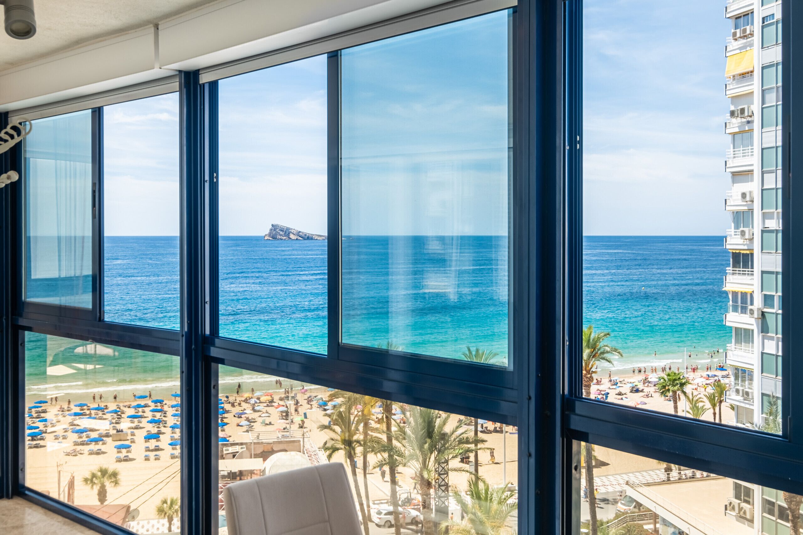 Un amplio ventanal con vista a una playa, palmeras y un edificio al otro lado del agua.