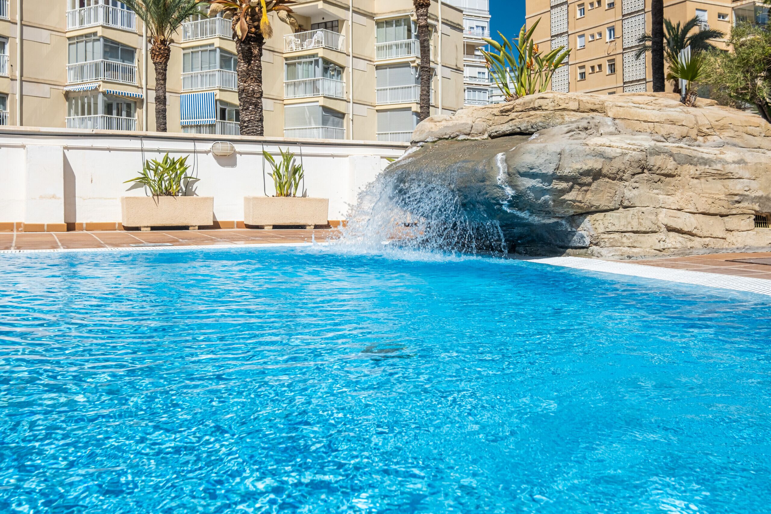 A swimming pool with clear blue water, a rock waterfall feature, and a view of a multi-story building with balconies and palm trees.