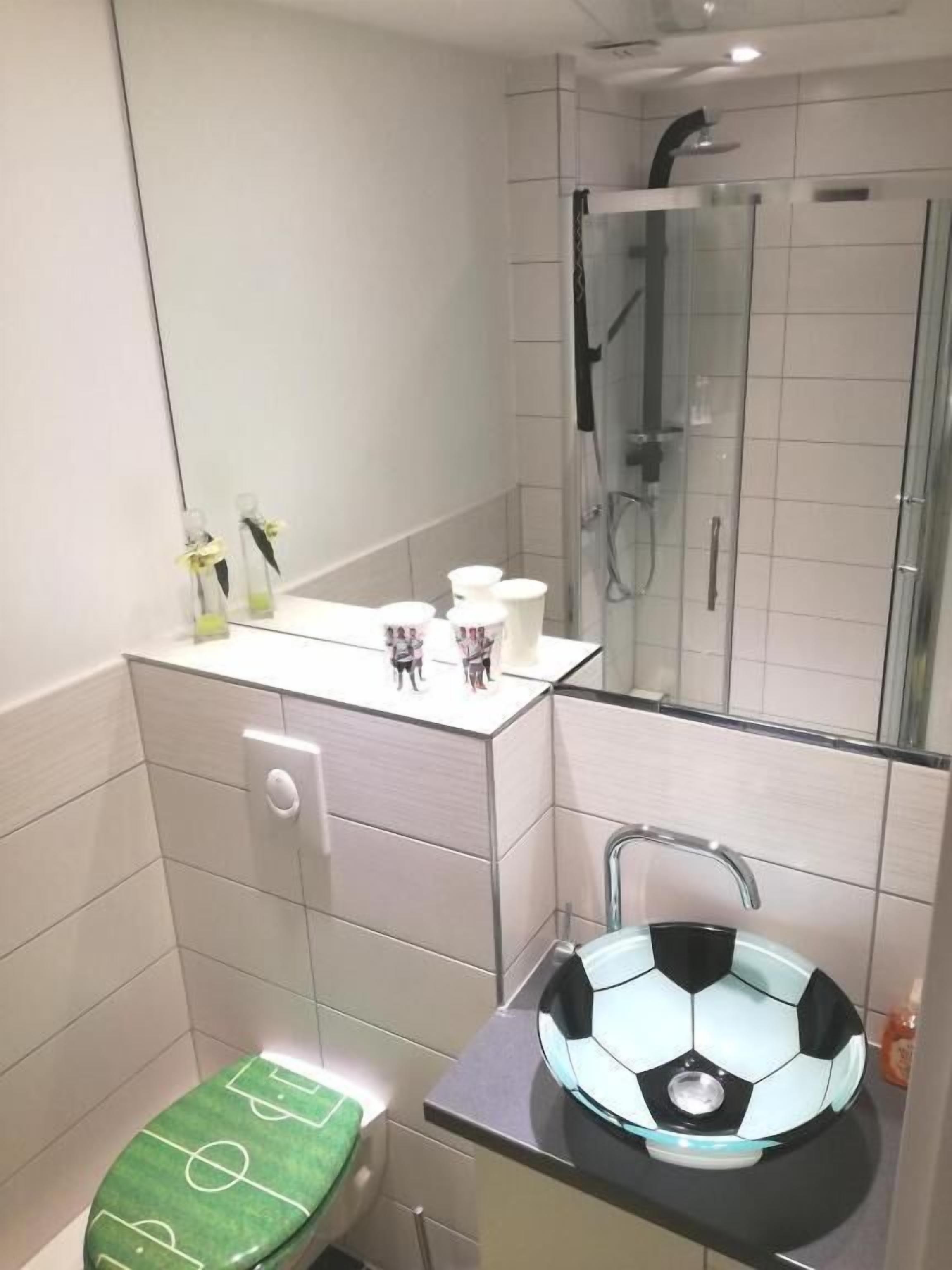 A modern bathroom with a glass shower enclosure, a toilet with a green seat, and a sink with a soccer ball design.