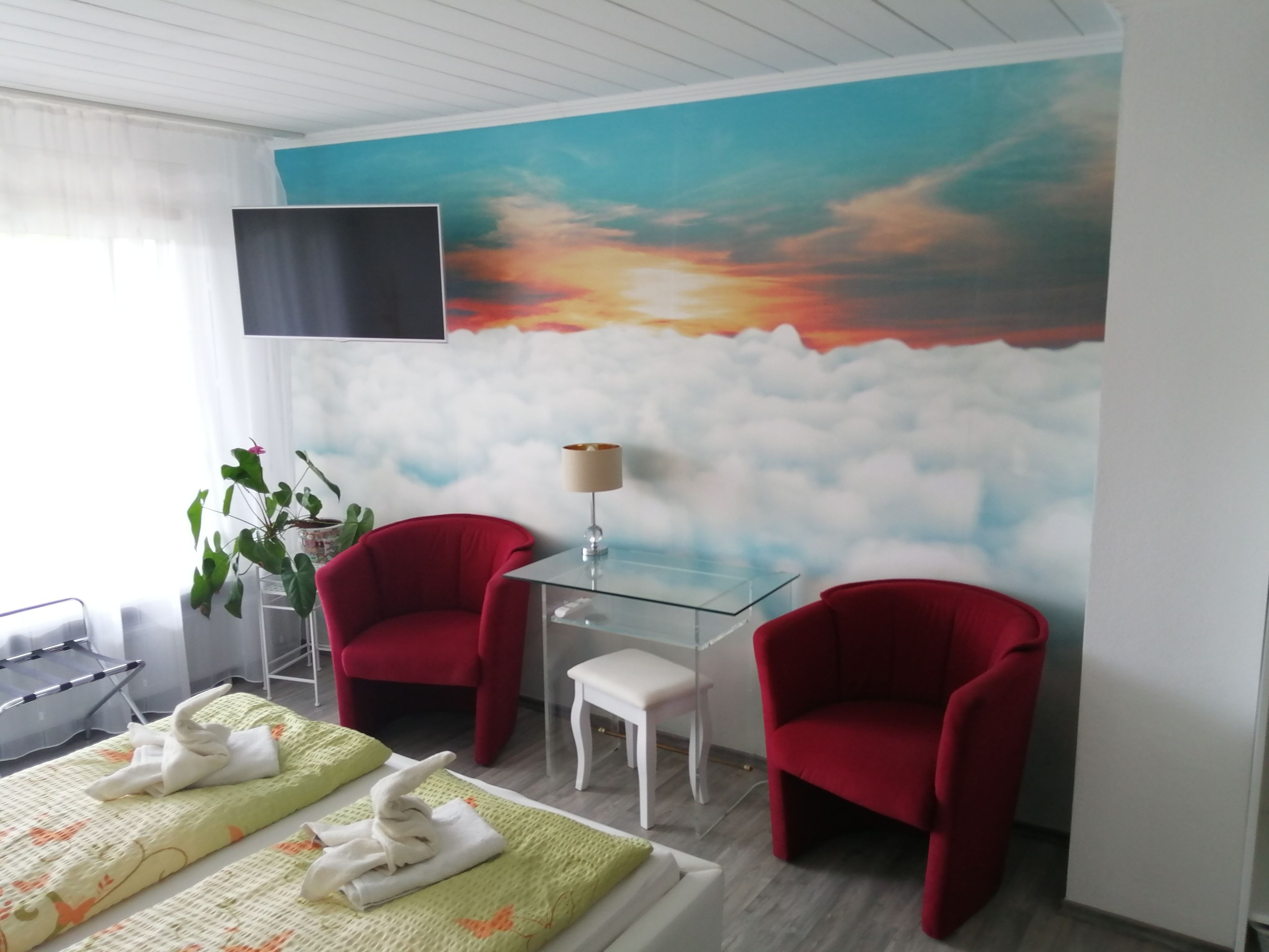 Una habitación de hotel con una cama, dos sillones rojos, una mesita con una lámpara y un gran mural de un atardecer en la pared.