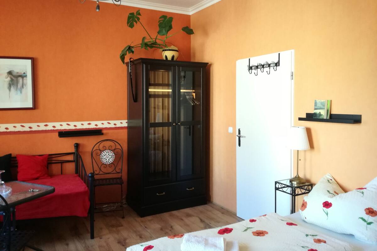 Quarto com parede laranja, um armário preto, cama com roupa de cama florida, uma cadeira vermelha e uma mesinha.