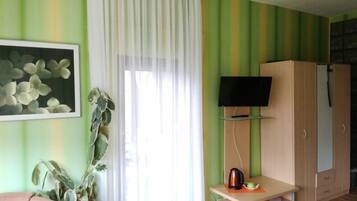 Une pièce avec un mur à rayures vertes, un rideau blanc, un téléviseur posé sur un meuble, une armoire, une plante et une petite table sur laquelle se trouvent une bouilloire et une tasse.