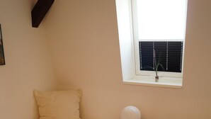 Quarto pequeno e simples, com duas camas, janela com persianas, mesa de cabeceira com abajur e piso de madeira.