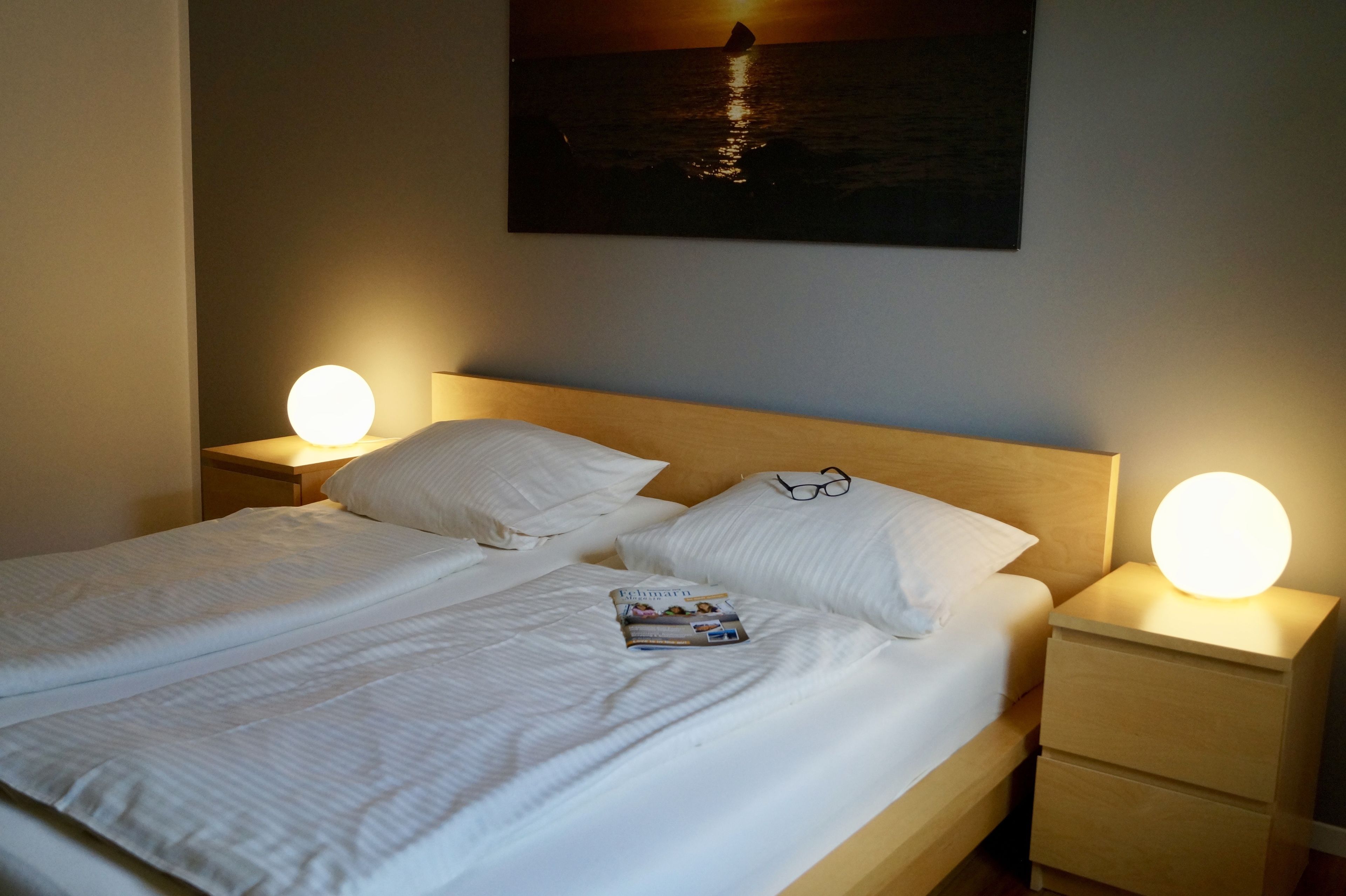 A neatly made bed with white linens, two pillows, and a magazine on the bed. There are two bedside lamps with round lights on, and a framed picture of a sunset over water on the wall above the bed.