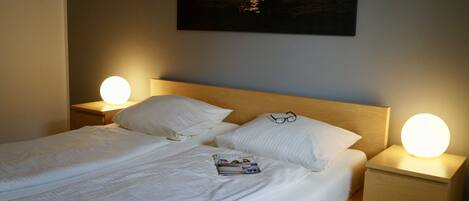 A neatly made bed with white linens, two pillows, and a magazine on the bed. There are two bedside lamps with round lights on, and a framed picture of a sunset over water on the wall above the bed.