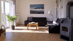 A living room with a sofa, a coffee table, a potted plant, a fireplace, and a large window. - Strandleben Fehmarn - Ferienhaus 1 (Fehmarn)