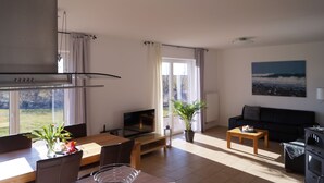 Ein modernes Wohnzimmer mit Essbereich, einer Couch, einem Fernseher und einem großen Fenster mit Vorhängen.