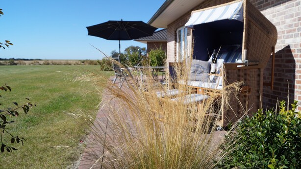 Eine Terrasse mit Korbstuhl und Sonnenschirm, ein Ziegelhaus und ein grünes Feld im Hintergrund.