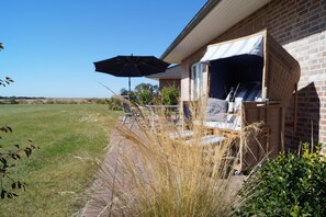 A patio area with a wicker chair and an umbrella, a brick house, and a grassy field in the background. - Strandleben Fehmarn - Ferienhaus 2 (Fehmarn)