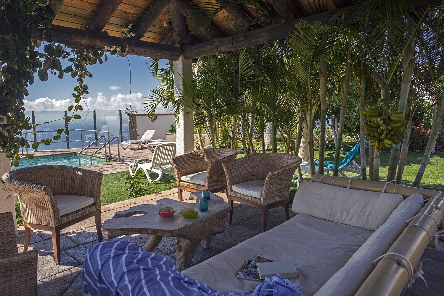 Un área de estar al aire libre con mobiliario de mimbre, una mesa de piedra y vistas a una piscina y palmeras.