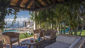 Area relax esterna coperta con mobili in vimini, un tavolo in pietra e vista su piscina e palme.