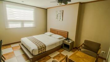 Una habitación de hotel con cama, mesita de noche, silla, una pequeña mesa, ventilador de techo y ventana con persianas.