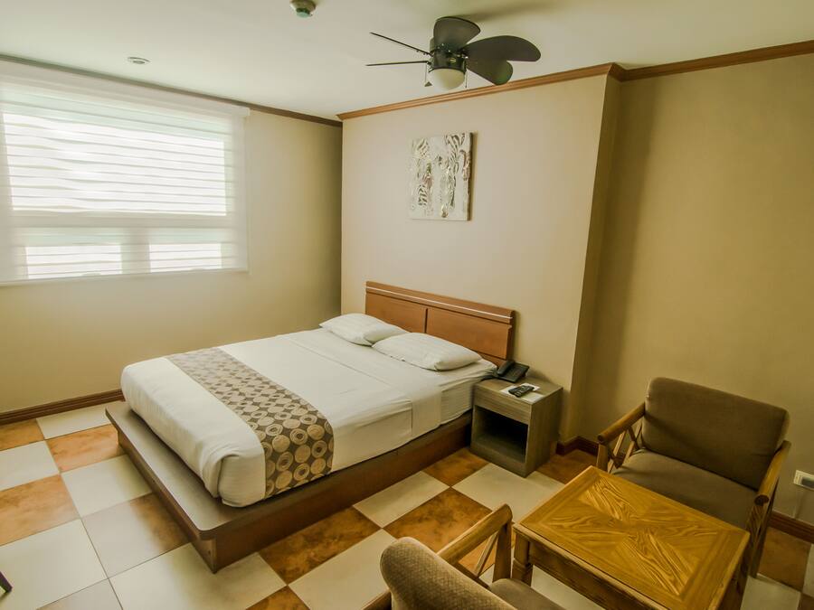 Una habitación de hotel con cama, mesita de noche, silla, una pequeña mesa, ventilador de techo y ventana con persianas.