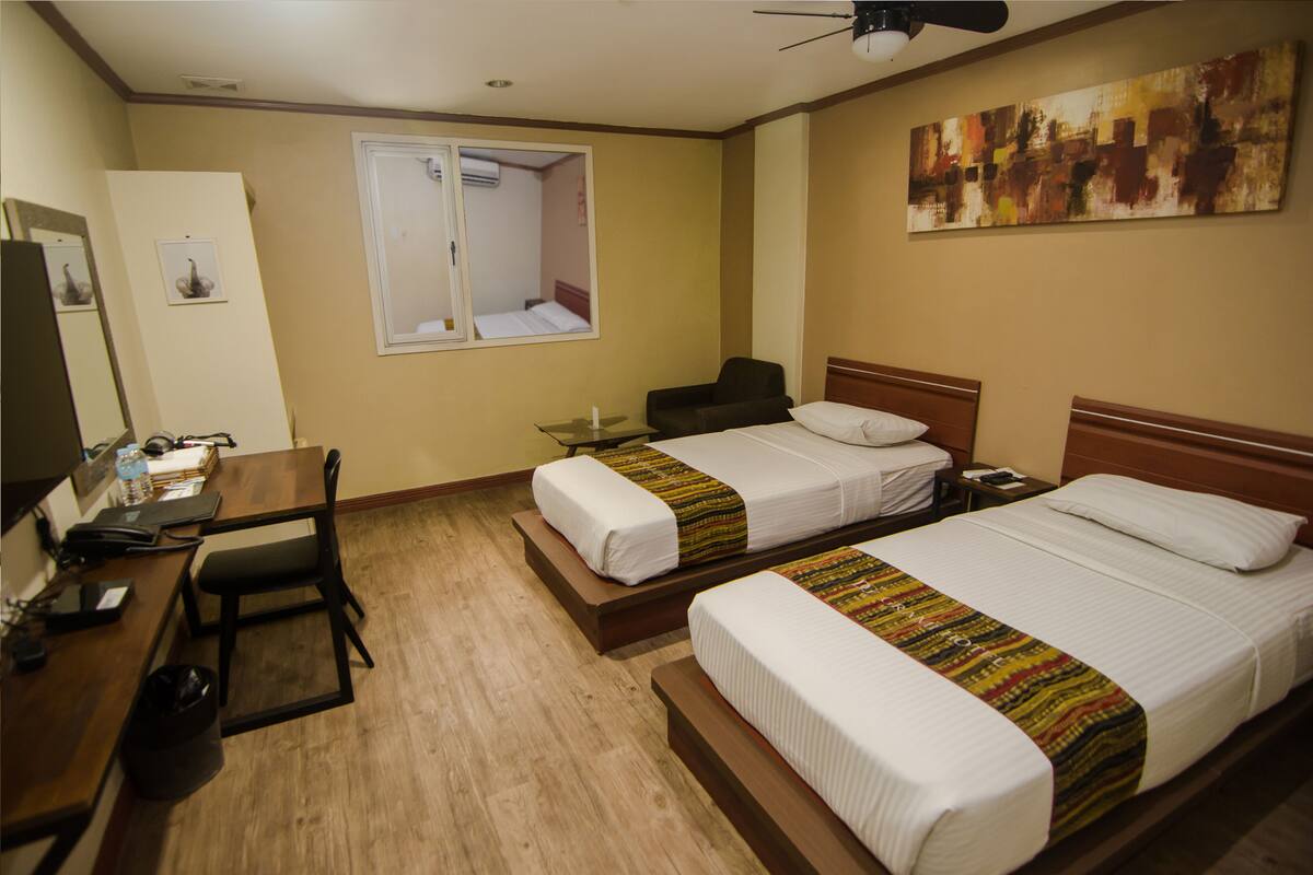 A hotel room with two beds, a desk, a chair, a TV, and a ceiling fan.
