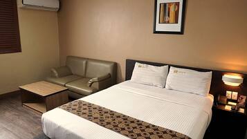 Habitación de hotel con cama, sofá, mesita, aire acondicionado, un cuadro en la pared y mesita de noche con lámpara y teléfono.
