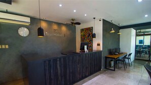 A hotel reception desk with a clock, a digital screen, and a seating area with chairs and a table. - DG Grami Hotel (Parañaque)