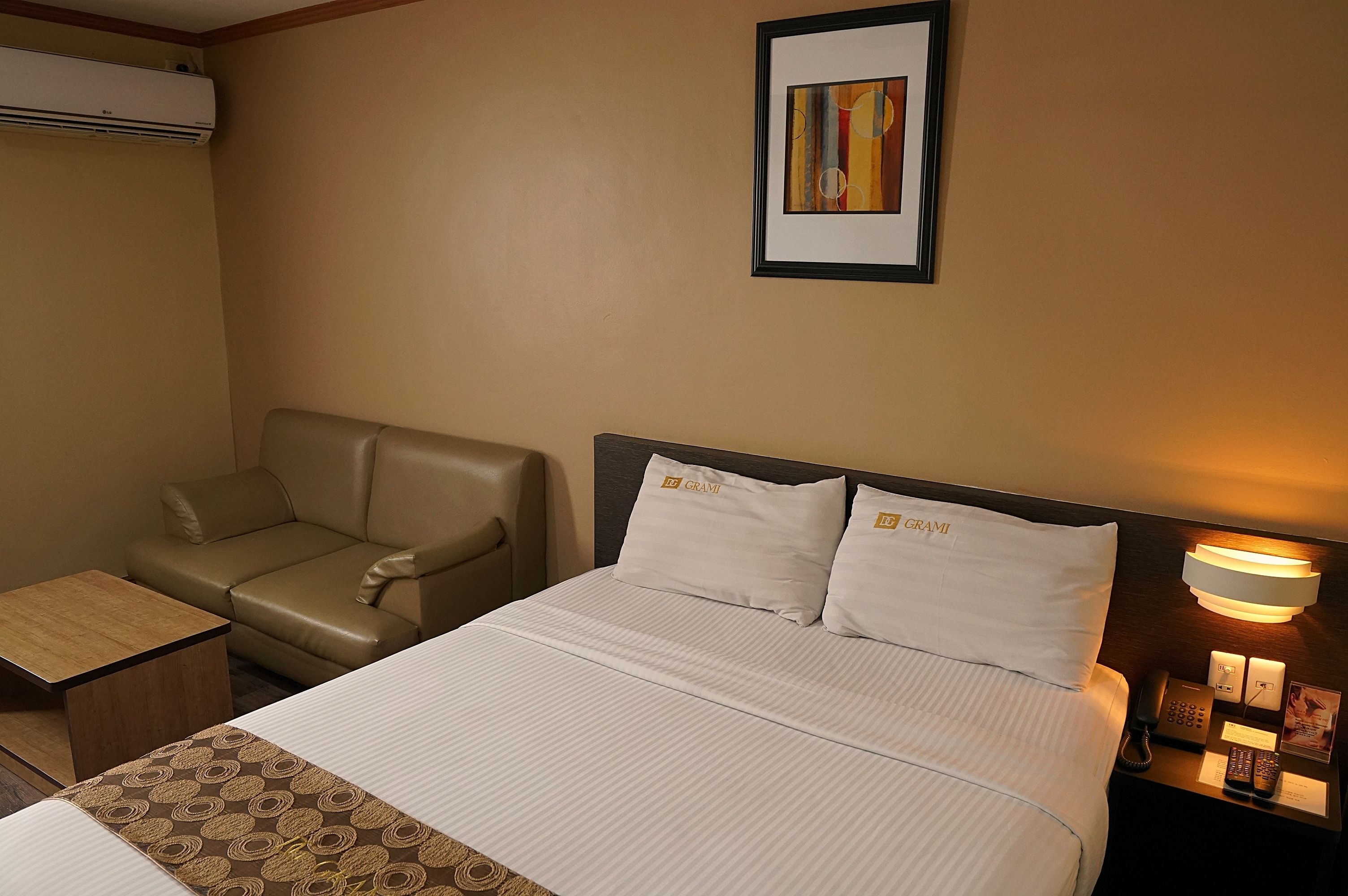 A hotel room with a bed, a sofa, a small table, a nightstand with a lamp, and a framed picture on the wall.