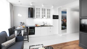 Een moderne keuken met witte kasten, een inbouwoven en een magnetron. Er is een eethoek met een tafel en stoelen, en uitzicht op een stadsgezicht door een geopende deur.