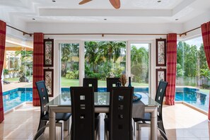 A dining area with a glass table and chairs, a ceiling fan, and a view of a swimming pool and garden. - Stunning Luxury Golf and Pool Villas (Pattaya)